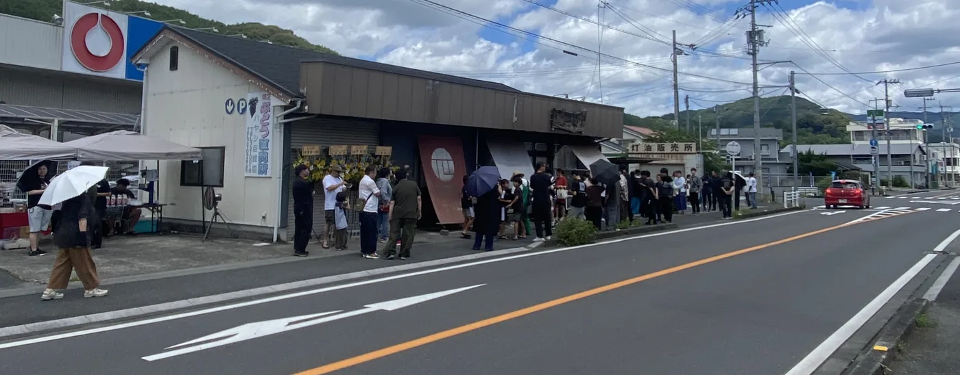 田舎の国道沿いにある麺や一心の前に、傘をさして順番を待つお客様の長い列ができている様子（愛媛県西予市野村町）